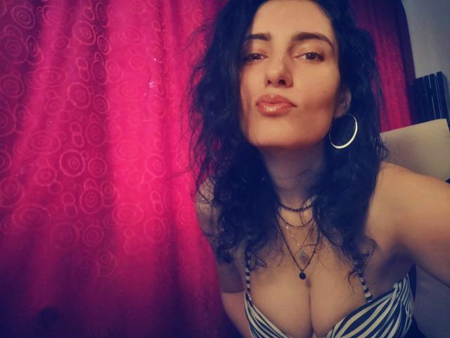 AddictedToUTS with curly black hair, hoop earrings, and striped top, pouting with a slight smile, showing her tits, wearing multiple necklaces, in a pink curtain-lit photo.