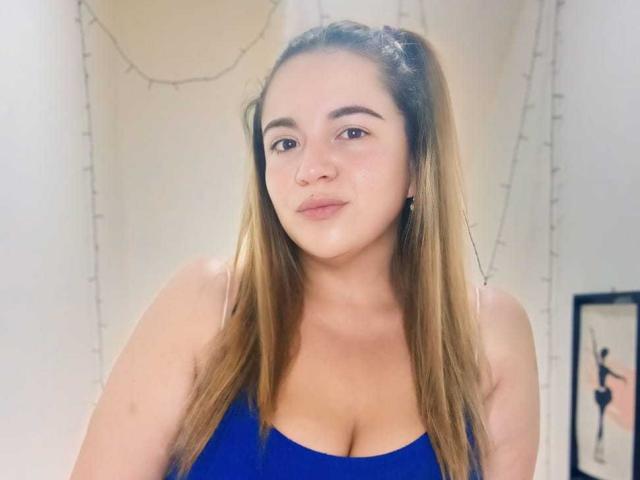 NaugthyCurvy, com cabelo castanho claro em maria-chiquinhas, vestindo uma blusa azul, mostrando os seios, tem uma expressão neutra e está em um quarto com iluminação suave..