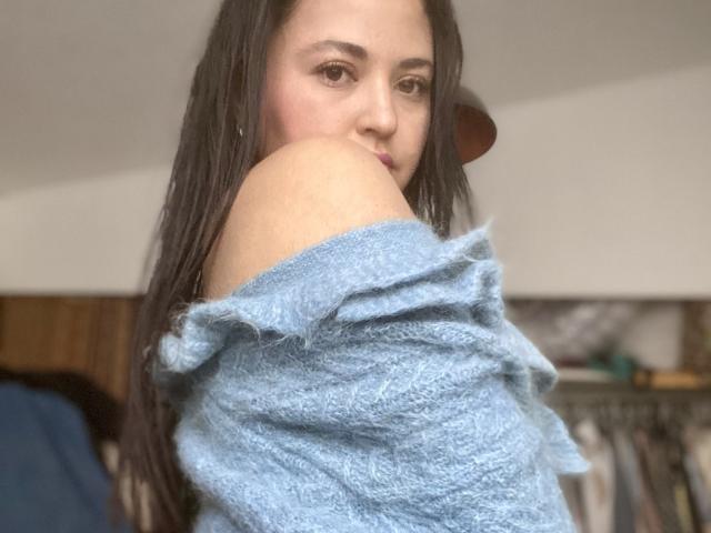 Hopeless with long dark hair, wearing a fluffy blue off-shoulder sweater, looking over her shoulder with a slight smile. Her shoulder is exposed, showing her bare skin.