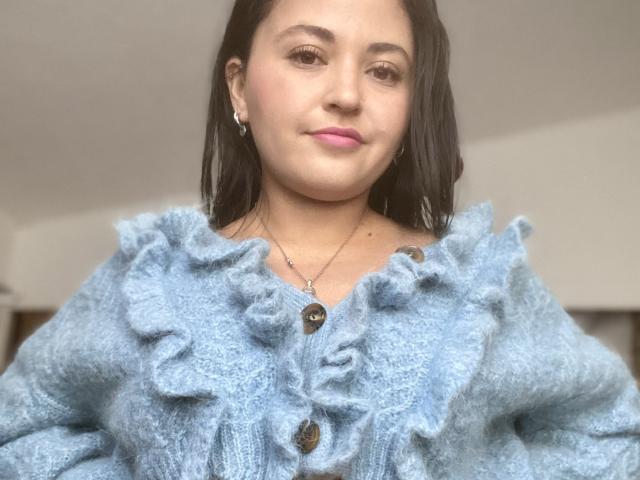 Hopeless with pale skin, pink lips, and black hair wearing a fluffy blue ruffle sweater, buttons, and a necklace, smiling slightly. Boobs are not visible.