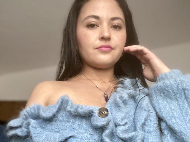 Hopeless with long black hair, pink lips, wearing a blue off-shoulder fluffy sweater, buttoned, and a necklace, looking slightly to the side, hand near face.