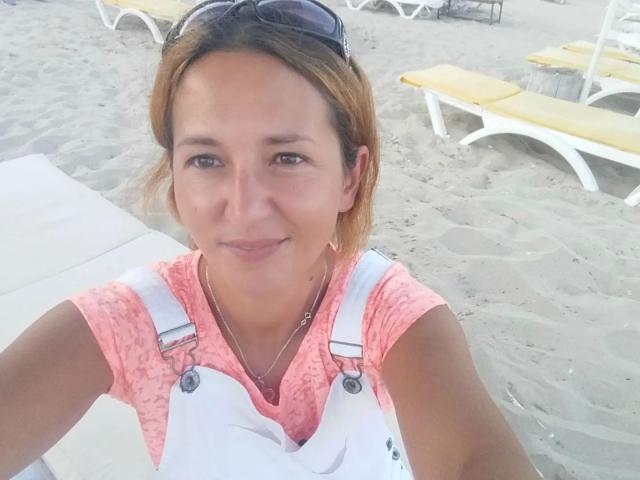 SubSandy, com cabelos castanhos curtos, vestindo uma blusa rosa de renda e macacão branco, tem seios pequenos e um sorriso discreto, e está em pé em uma praia arenosa com espreguiçadeiras amarelas..