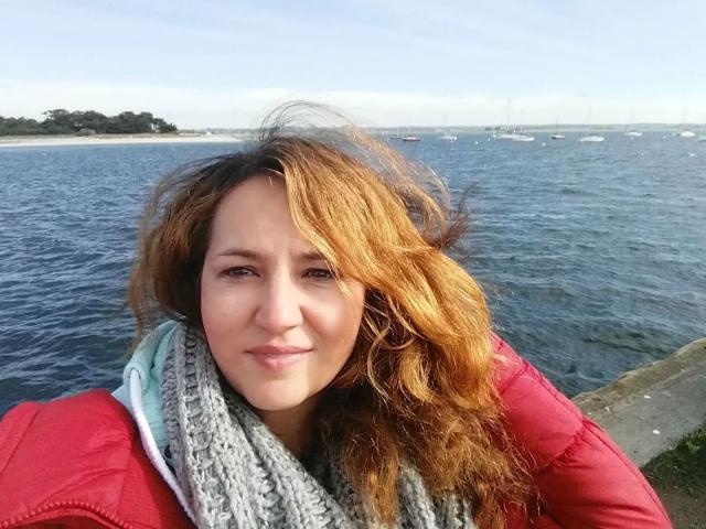 SubSandy-hot a les cheveux ondulés, brun-orangés, une peau claire et porte une veste rouge avec une écharpe grise. Elle sourit, et se tient près d'une étendue d'eau bleue avec des bateaux en arrière-plan..
