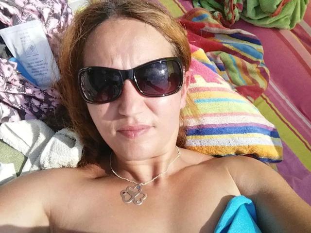 SubSandy - morena, con piel bronceada, cabello castaño, gafas de sol negras y un collar, desnuda, recostada sobre una toalla de colores, mostrando sus pequeños senos y un paño azul en su mano..