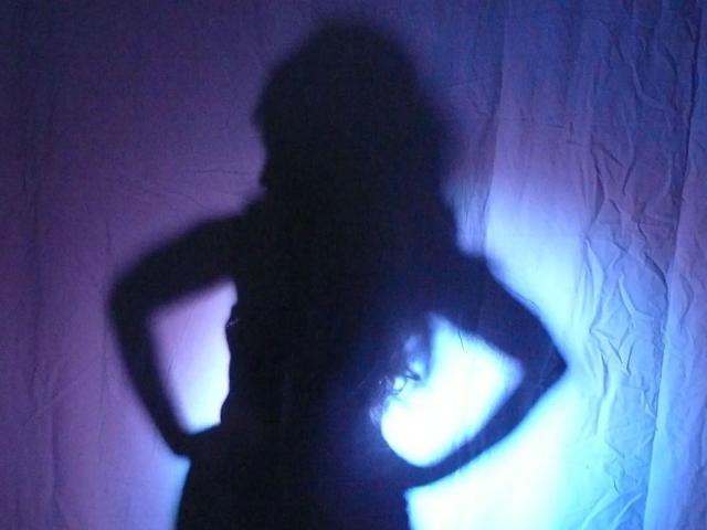 Silhouette of SubSandy-hot with hands on hips, dark shadow with blue light highlighting the curve of her ass and long wavy hair against a wrinkled, purple background.