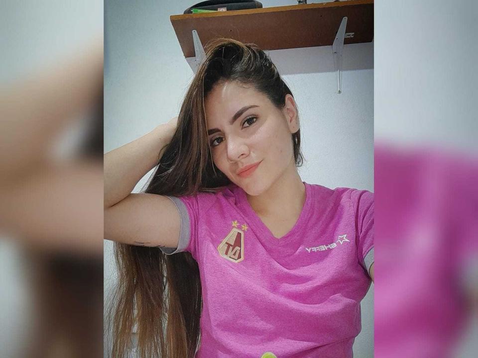 ValeriaCoox with long brown hair, wearing a pink sports shirt, hand in hair, smiling, small nose ring, white wall, shelf with wooden top in background.