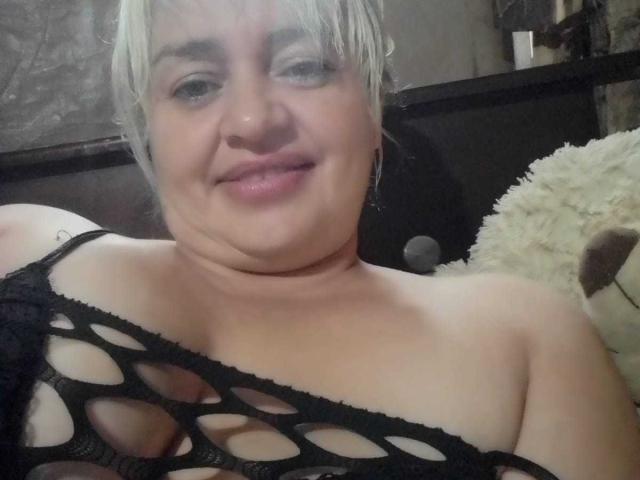 SalomeAriza has short blonde hair, a slight smile, and is wearing a black fishnet top that reveals her large, soft tits. A fluffy white toy is behind her.