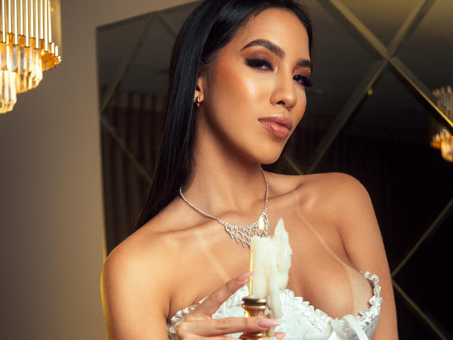 Kylie White, con piel morena, cabello negro y labios brillantes, sosteniendo un plumero blanco cerca de sus pequeños senos, vistiendo un collar de plata y una blusa blanca de encaje..