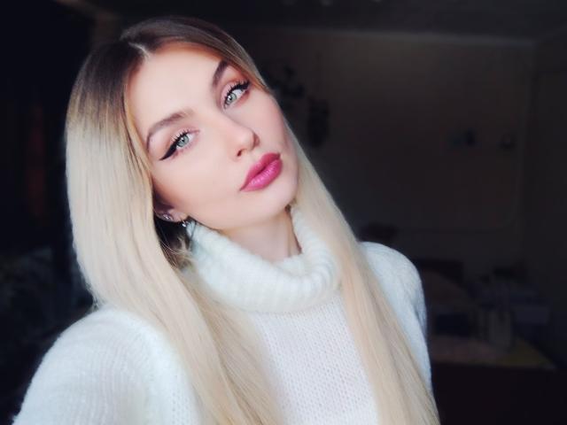 EvaLoona, con cabello rubio largo, ojos verdes, lápiz labial rosa y un suéter blanco de cuello alto, mirando ligeramente hacia arriba. Sus pechos son pequeños, su rostro es bonito y es atractiva..