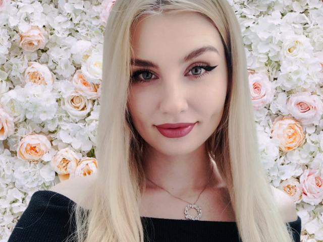 EvaLoona with blonde hair, dark lipstick, and black off-shoulder top, wearing a heart necklace, has cute ass-adjacent face, flirty eyes, and a subtle smile.