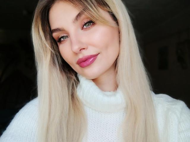 EvaLoona, con i capelli biondi, gli occhi verdi, il rossetto rosa, il maglione bianco a collo alto e un sorriso dolce, che mette in mostra il suo bel viso e il suo sedere carino in questa foto..