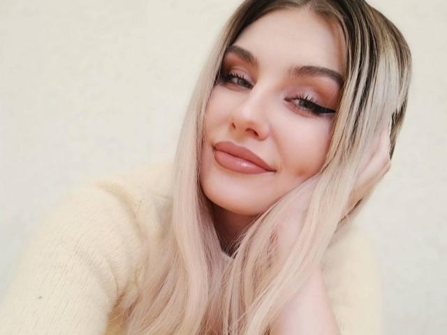 EvaLoona ha lunghi capelli biondi, labbra piene rosa e ciglia folte, indossa un maglione giallo, sorride con una mano tra i capelli, appare carina e sensuale..