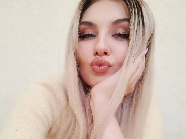 EvaLoona, con labios rosados y fruncidos, cabello rubio largo y pestañas largas, vestida con una blusa de punto color crema, con su mano tocando suavemente su mejilla. Piel suave y pálida..