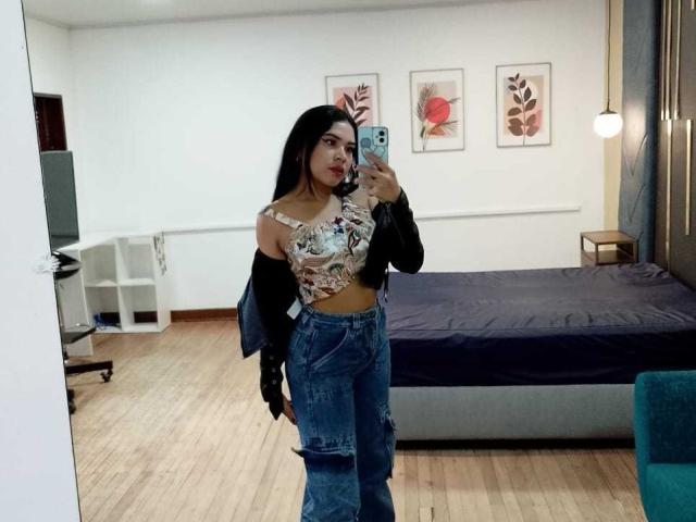 OliviGrace with long black hair, wearing a patterned crop top, denim skirt, black jacket, and black gloves, taking a mirror selfie in a bedroom. Boobs visible.