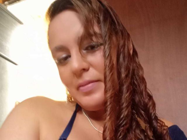 AmandaHotL with wet, brown, curly hair, blue halter top, and pink lipstick, looking down, soft smile, bare shoulders, small silver necklace, light skin, warm lighting.