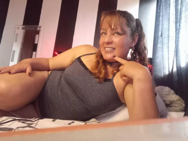 AmandaHotL with red hair in a ponytail, wearing a gray tank top, lying on a bed, smiling, touching her chin with her finger, showing her tits.