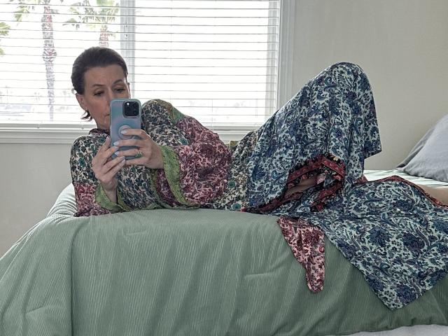 SkyChick lying on a bed, wearing a colorful floral dress, holding a phone, taking a selfie, with her ass and tits partially visible, in a bright room.