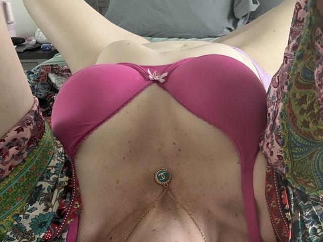 SkyChick lying on a bed, pink bra covering her tits, purple panties, gold necklace with green gem, floral robe, legs spread, soft light, pale skin, relaxed.