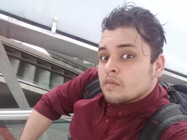 Bigchef with dark, wet hair, brown eyes, and a slight beard, wearing a red shirt, looking at the camera with a neutral expression, and a black backpack.