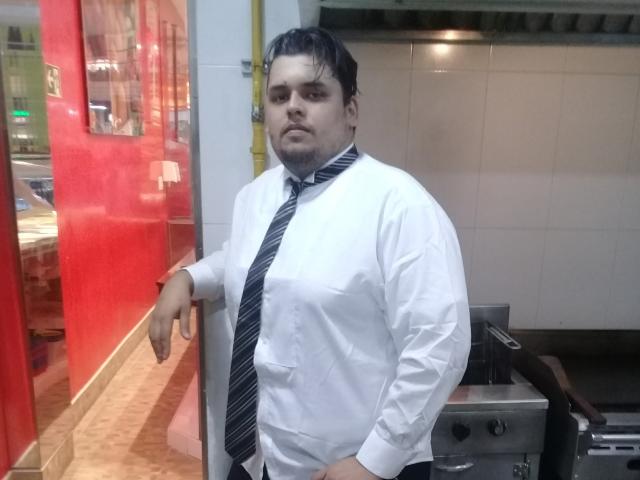 Bigchef is a chubby dude with black hair, wearing a white shirt and loose black tie, standing with a bored expression, leaning against a red wall.