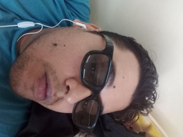 Bigchef has dark glasses, a teal shirt, and white earbuds. He's looking at the camera with a slightly open mouth, showing a few moles on his cheek.