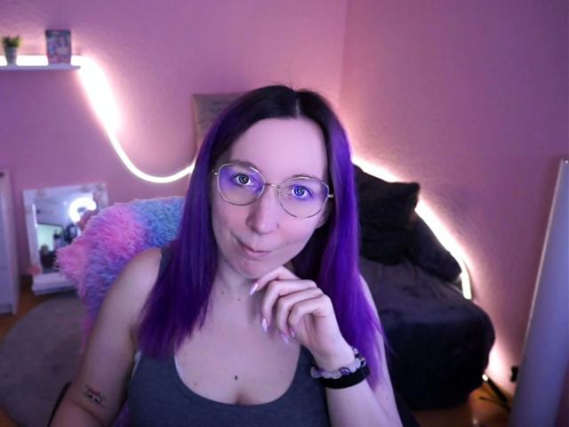 JustKerry-hot with purple hair, glasses, and a grey tank top, pouting with her finger to her lips, showing her tits and a tattoo on her left arm.