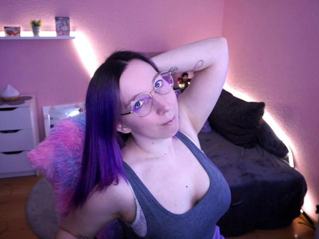 JustKerry-hot with purple hair, glasses, gray tank top, showing her tits, arm raised, tattoo on arm, in pink-lit room, sitting on chair with pink and blue fuzzy pillow.