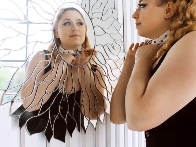 JessikaDaniels with wavy blonde hair, black dress, white polka dot choker, looking at sun-shaped mirror with her reflection, hands near neck, in bright room.