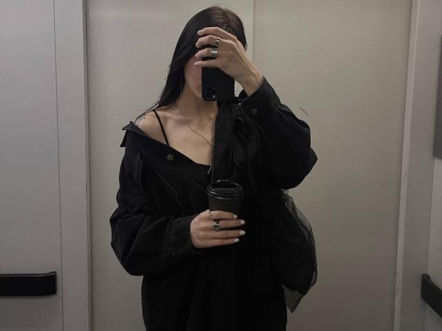 Tsukerberg with long black hair, wearing a black off-shoulder top and jacket, holding a coffee cup, taking a mirror selfie with her phone, showing her tits slightly.