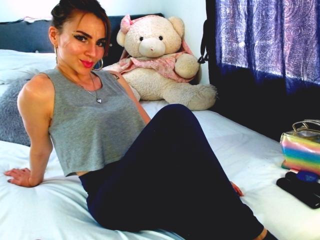 EmelySofia with tits in a gray crop top and black pants, sitting on a bed, smiling, with a large white stuffed toy and a rainbow purse nearby.