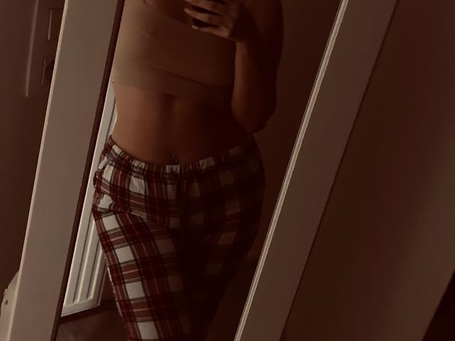 A mirror selfie of SweetHottBabe69 with a flat stomach, tan bra, and red plaid pajama pants, showing a belly button piercing, in dim lighting.