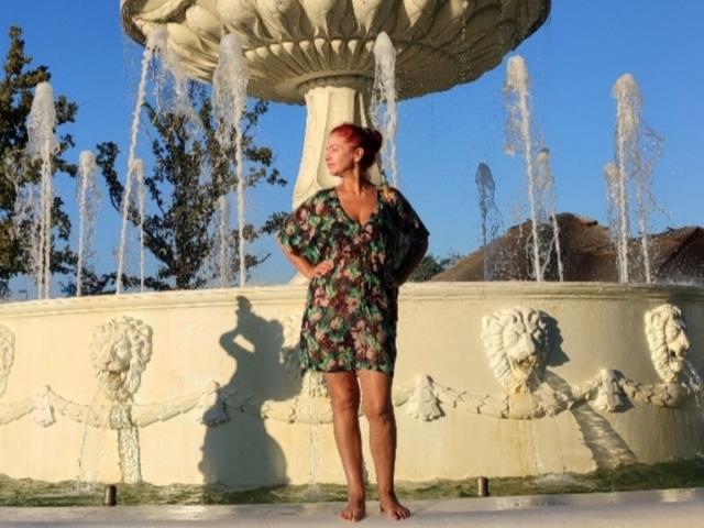 TaisieaViolet stands barefoot in front of a fountain, wearing a green floral dress, hands on hips, water spraying from the top, her shadow on the fountain's side.