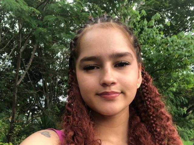 MelodyMansson has curly red hair, brown eyes, and a small tattoo on her right shoulder. She's smiling with light makeup and wearing a pink top.