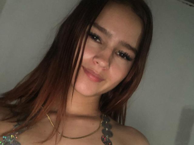 Melody Mansson tem cabelos castanho-claros, cílios longos e um sorriso discreto. Ela está usando um colar com contas coloridas, mostrando seus ombros e um leve decote..