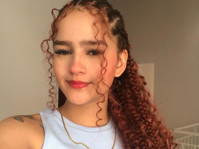 Melody Mansson a les cheveux roux bouclés, une peau claire et du rouge à lèvres rouge. Elle sourit, ses lèvres rouges sont visibles, elle porte une chemise bleu clair et a un petit tatouage sur son épaule gauche..