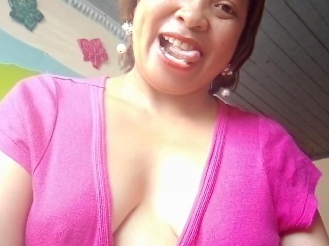 Rahel69 with a playful expression, tongue out, wearing a pink V-neck shirt that shows her big, round boobs. She has brown hair, pearl earrings, and a fair skin tone.