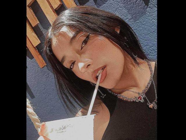 SalomeMille, con cabello negro, sorbiendo con una pajita, sosteniendo una taza blanca, vistiendo una blusa negra y un collar con una gema roja, con una pared azul soleada de fondo..