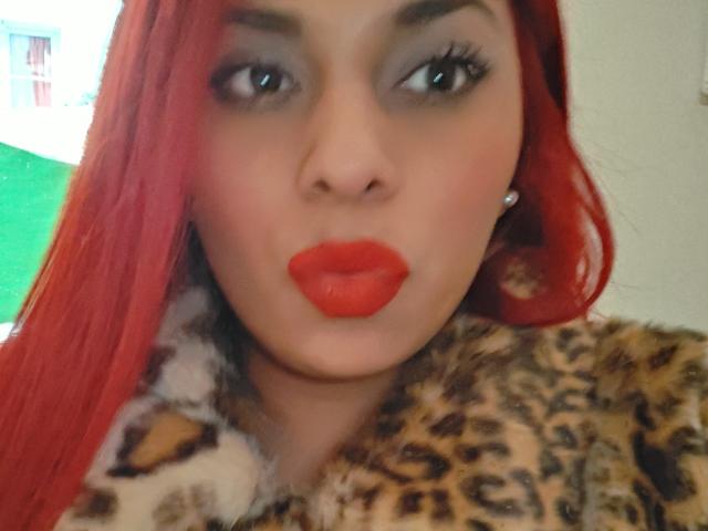 Chili avec du rouge à lèvres rouge vif et des cheveux rouges, portant une fourrure imprimée léopard, des lèvres moites et regardant directement la caméra.