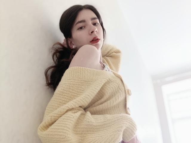 WollyMolly with dark hair, pale skin, and red lips, wearing a yellow knitted sweater, has one shoulder exposed, looking down with a slightly open mouth.