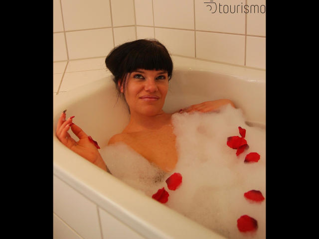 Jenny-Sun, with dark hair in a bun, is in a white bathtub filled with bubbles and red rose petals, smiling at the camera, showing her bare tits.