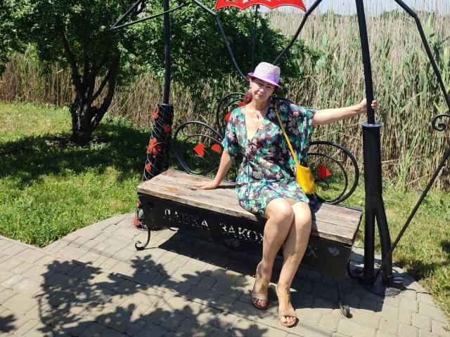 TaisieaViolet, with tits visible in a floral dress, sits on a wooden bench, her ass slightly raised, wearing a pink hat, yellow bag, and sandals, in a sunny park.