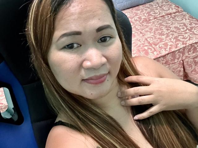 SimplyPinay has light brown skin, long brown hair, and is smiling slightly with her hand on her neck. She's wearing a black top, showing her tits.