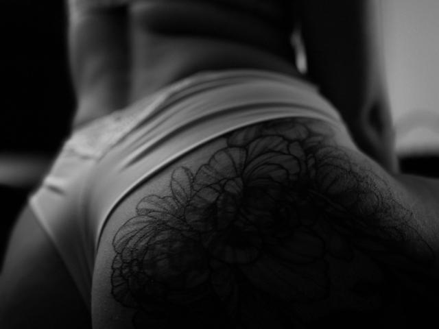 Black-and-white photo of CoreMelissa's lower back, showing a large, intricate tattoo of dark, floral patterns over her hairy ass in tight, white underwear.