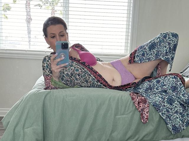 SkyChick lying on a green bed, wearing a floral robe, pink bra, and lavender panties, taking a mirror selfie with a blue phone, showing her tits and pussy.