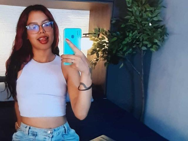 RouseGilmore with red hair, glasses, and white crop top, sticking tongue out, holding blue phone, wearing light blue jeans, standing in front of a green plant.
