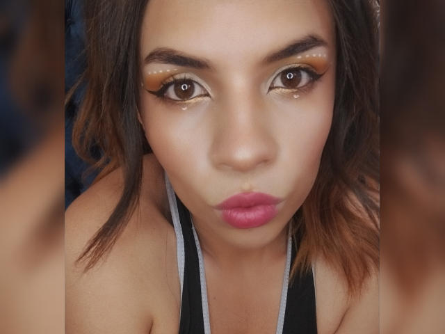 VioletaStone's face, with shiny gold eyeshadow, brown eyes, pink lips, and wavy brown hair, wearing a black halter top, looking straight into the camera with a pout.