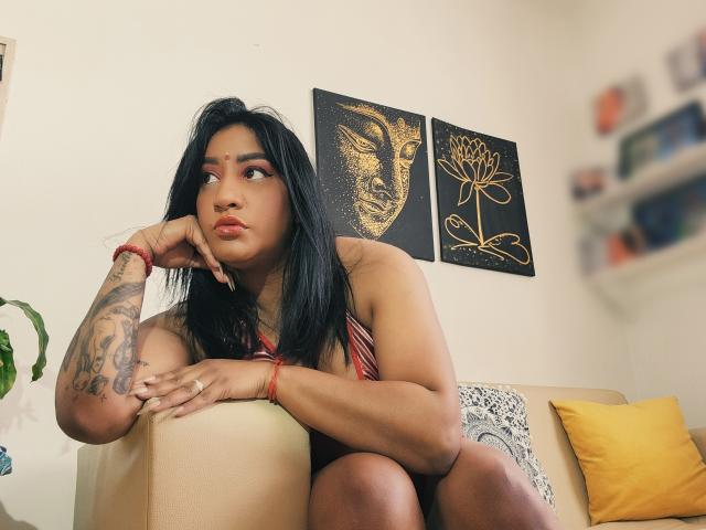 Nahirr, with black hair, sits on a beige sofa, her tattooed arm resting on her chin, wearing a red and black striped top, looking to the side.