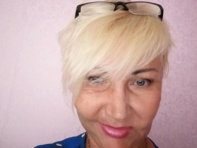 HellyHots-hot has short, bleached blonde hair, blue eyes, and pink lipstick. Black-framed glasses are on top of her head. She's wearing a blue top with white polka dots.