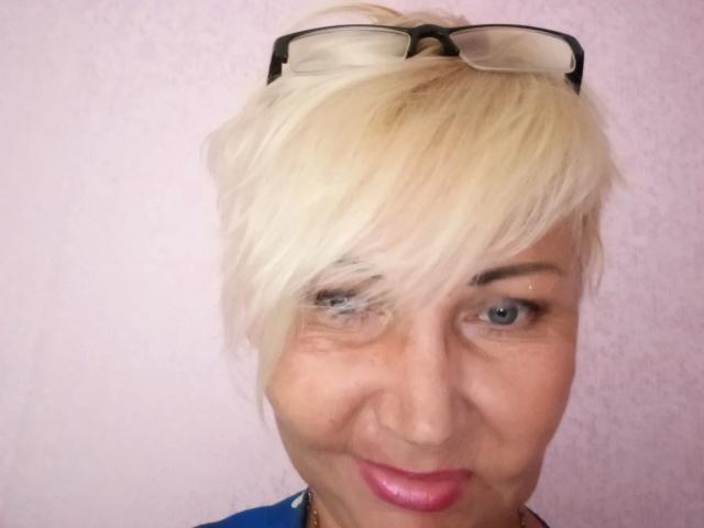 HellyHots-hot has short, messy blonde hair with glasses on top, blue eyes, a slight smile, and pink lipstick. She's wearing a blue shirt.