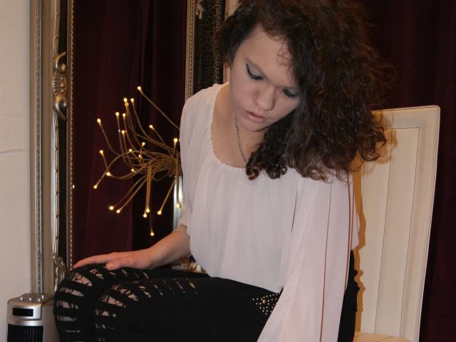 Wildandsexy25-hot with curly brown hair, white blouse, and black ripped pants, sitting on a chair, looking down, with a reflective mirror and decorative lamp in the background.